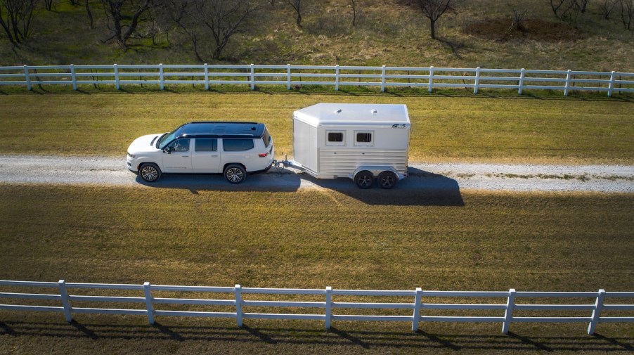 Overhead view of a white Grand Wagoneer SUV towing a horse trailer down a driveway, white fences visible on both sides.