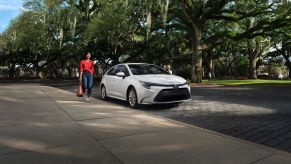 Woman in red holding a coffee walking toward a new white 2023 Toyota Corolla sedan parked on a street