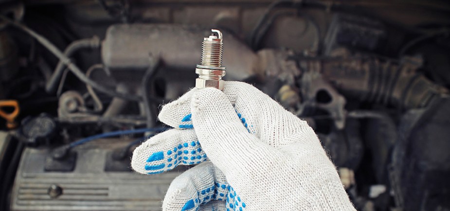 A gloved mechanic's hand holds a brand new spark plug above an older engine.