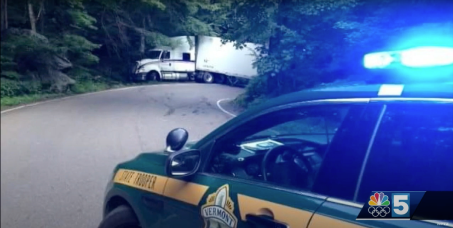 Tractor trailer stuck in the Stowe Smuggler's Notch mountain pass with a Vermont state trooper's car visible in the foreground.