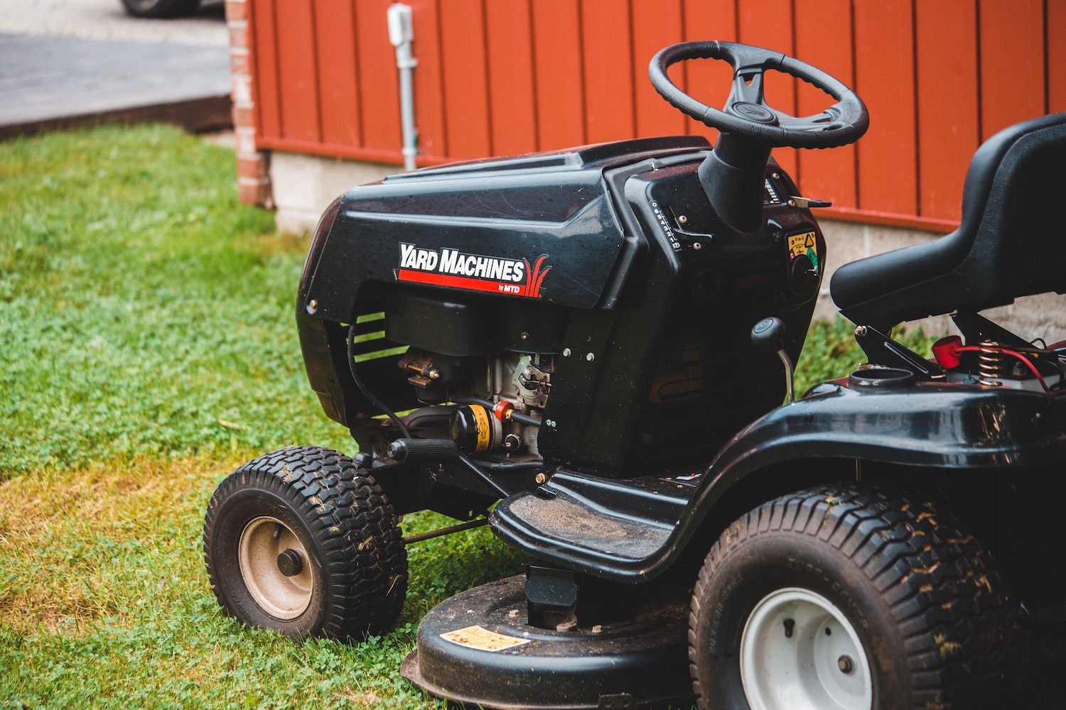 Black lawn tractor parked on a yard by a red shed wall.