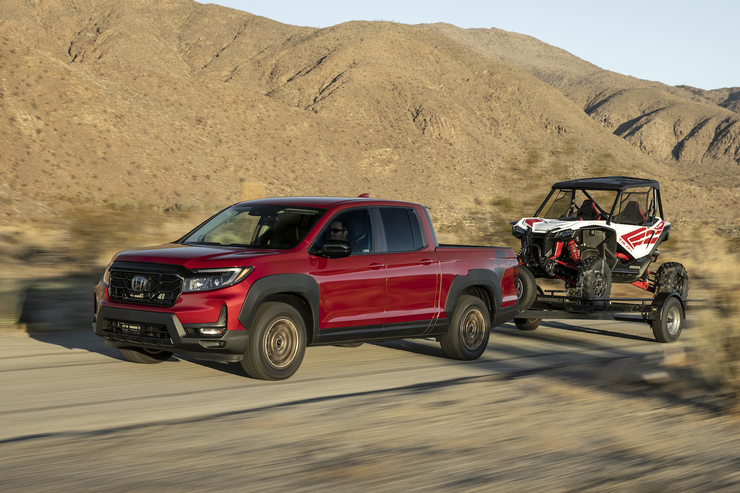 Red Honda Ridgeline pickup truck demonstrating its towing capacity by trailering in the desert, mountains visible in the background.