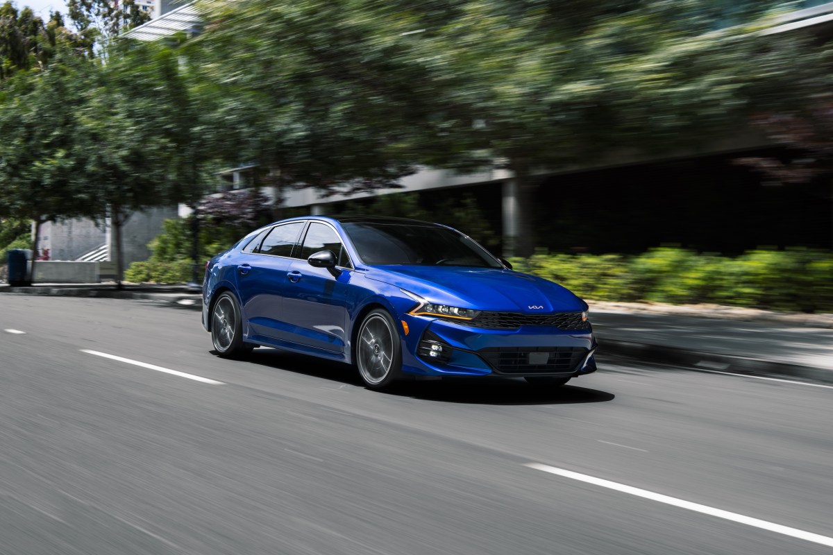 A blue 2022 Kia K5 driving down a tree-lined city street