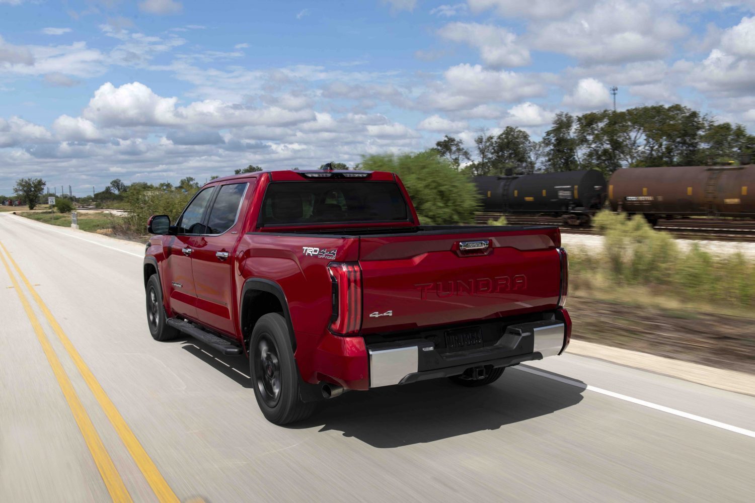 Red Toyota Tundra 4x4 pickup truck driving away down a country road, a train visible in the background.