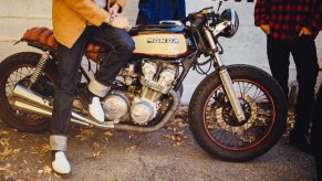 Customized cafe racer motorcycle parked by a brick wall, its rider sitting atop its seat, two other people's legs visible nearby.