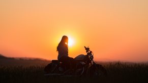 Woman sitting atop a Harley Davidson motorcycle while watching the sun set, a grassy field visible in the background.