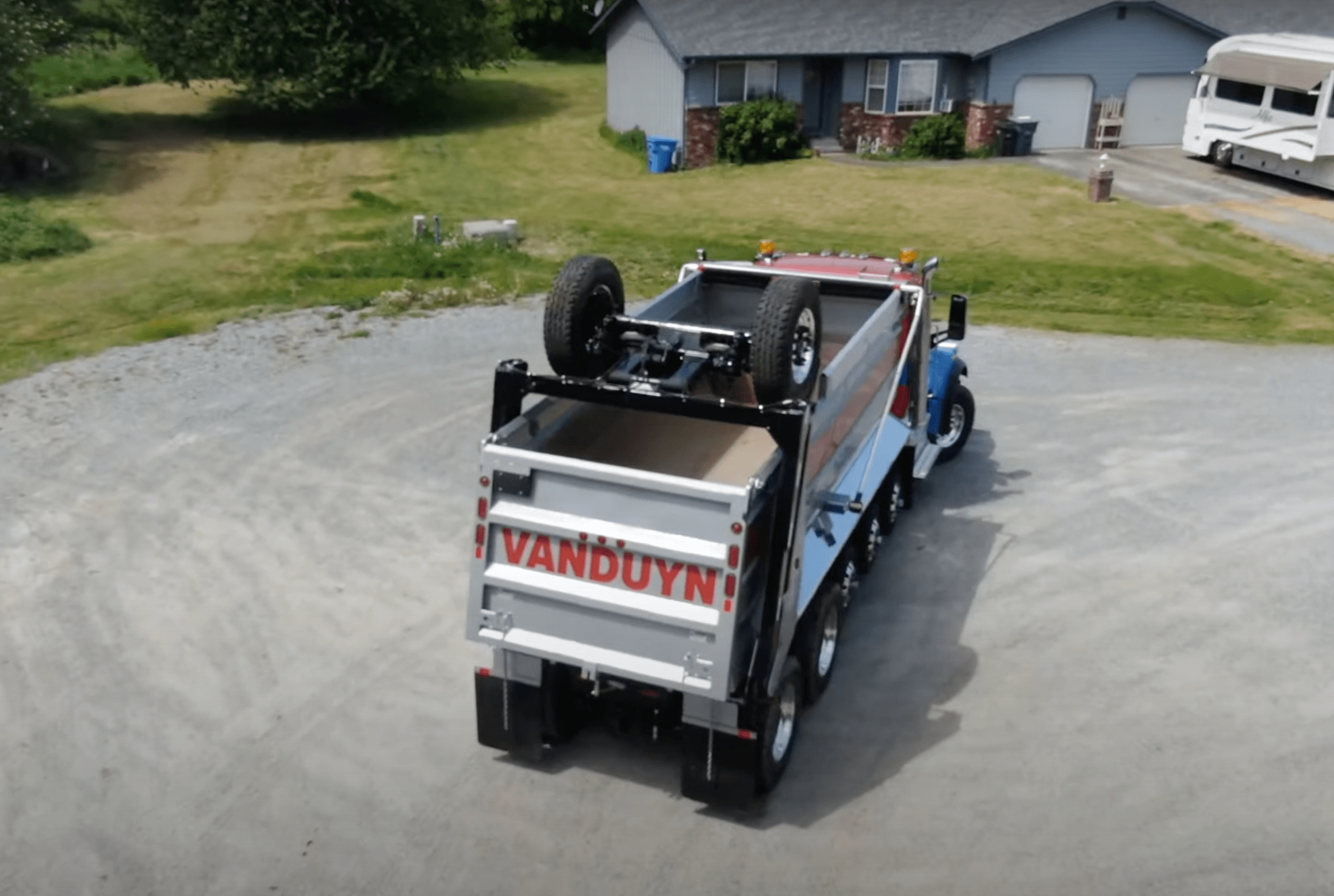 A dump truck with a rooftop drop axle and two wheels, a suburban home visible in the background.