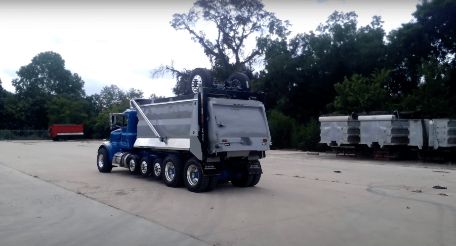 Blue dump truck with a rooftop drop axle and two wheels, a parking lot visible in the background.