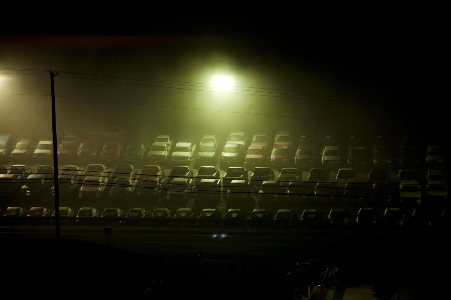 Rows of full-size pickup trucks parked under street lamps at a dealership, after dark.