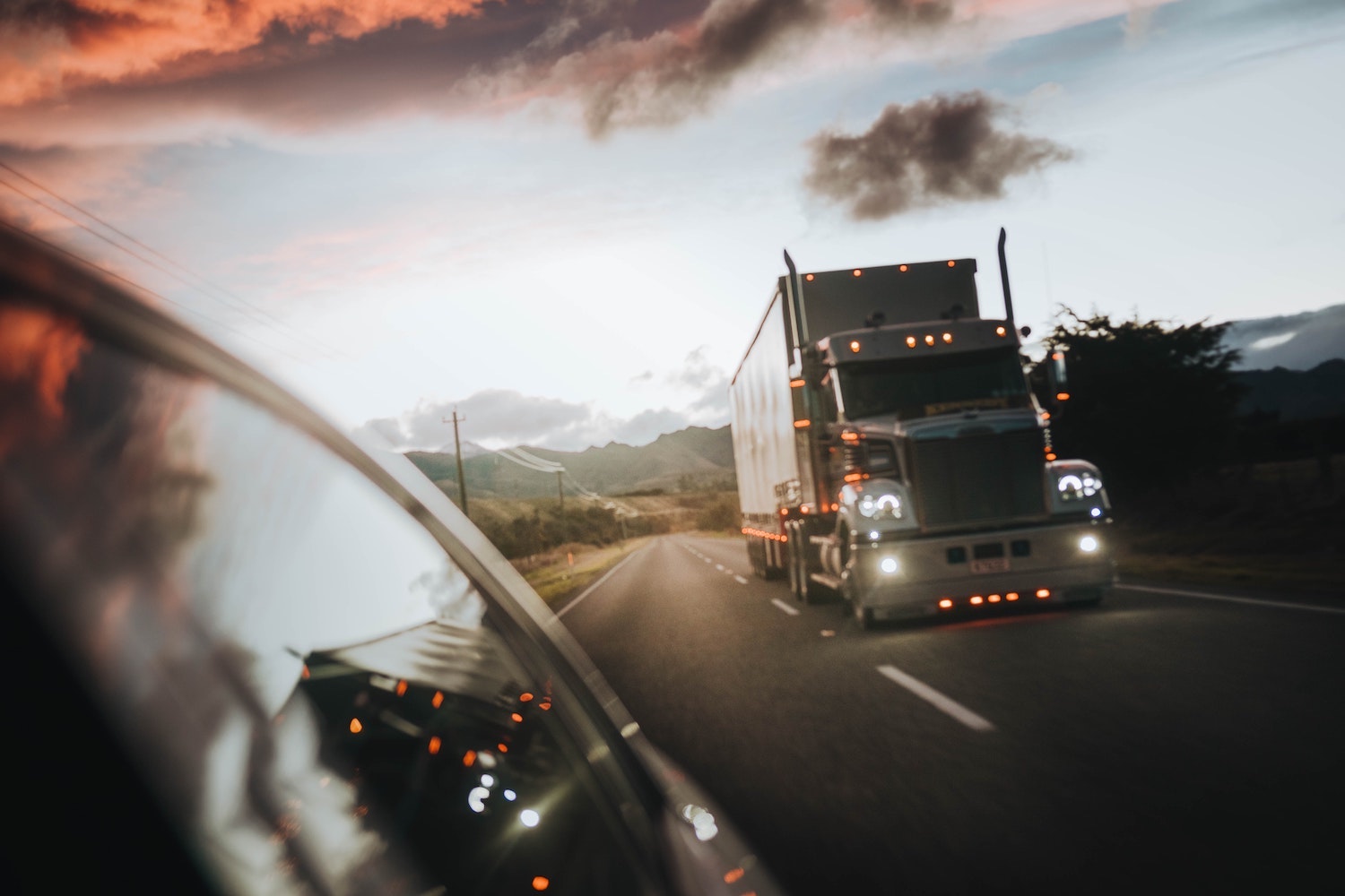 A tractor trailer driving down a country road at dusk with its amber marker lights glowing.