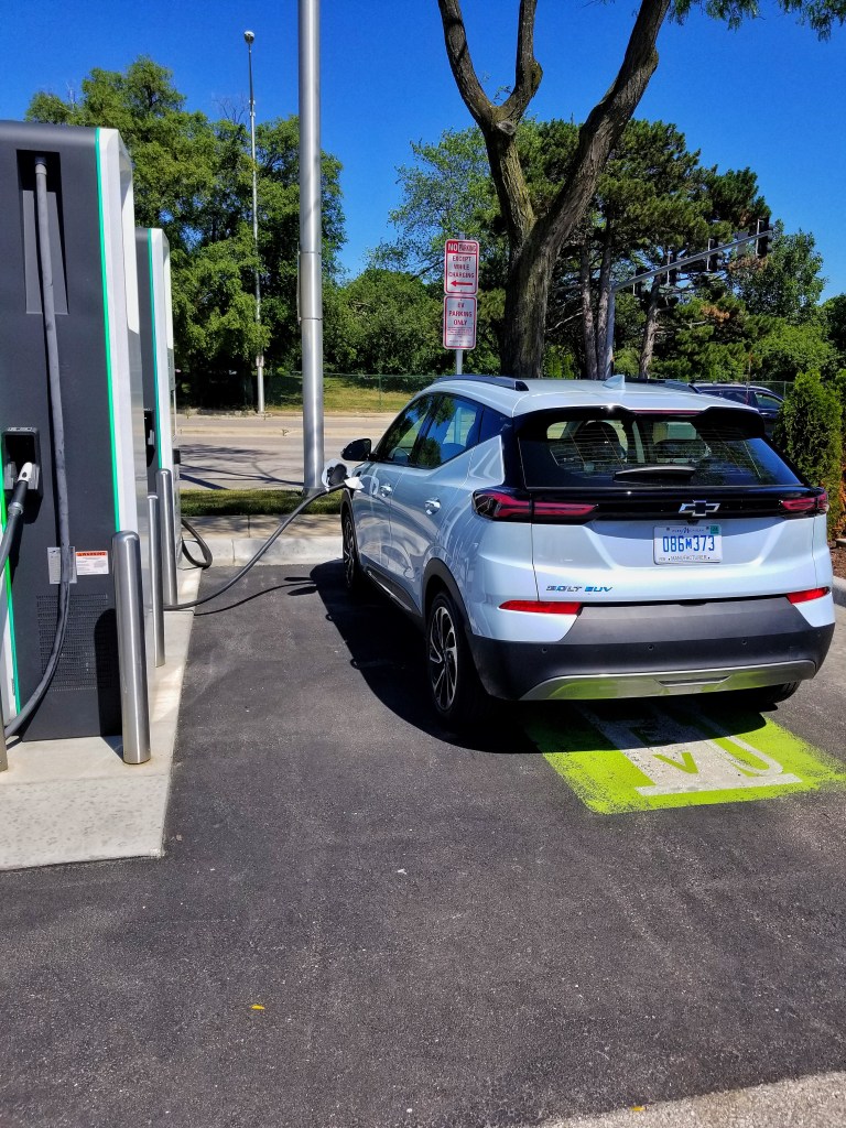 The side 3/4 view of a light-blue 2022 Chevrolet Bolt EUV parked by a white-and-gray Electrify America charging station in a shopping mall parking lot