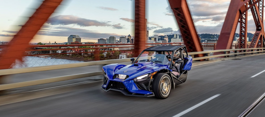 A blue Polaris Slingshot drives over a bridge.