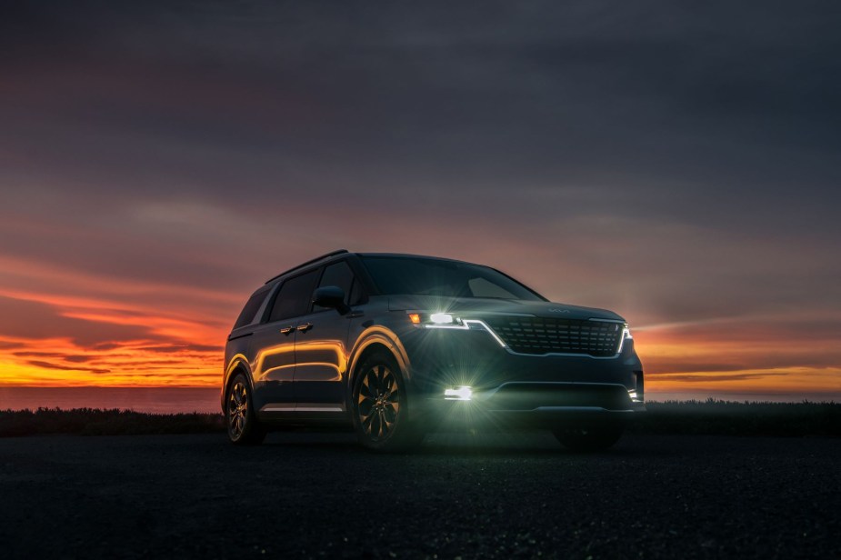 A 2023 Kia Carnival minivan model parked in a field at sunset with front headlights turned on