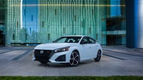 A white 2023 Nissan Altima, a good family car, sitting in front of a green lined wall on pavement.