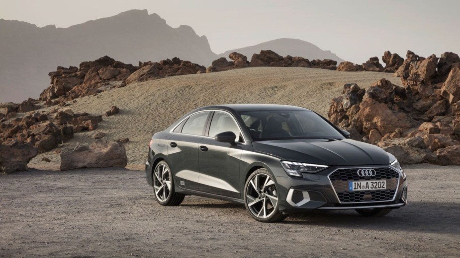 A dark gray Audi A3 sedan luxury car model parked on a dirt plain near a sand dune and rocks