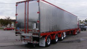 A refrigerated semi truck trailer with an open rear vent door, the truck tractor and a street visible in the background.