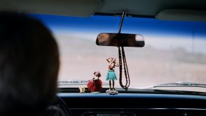 Image of two hula girl figurines on an old cars dashboard, the road beyond the windshield is out of focus and blurry.