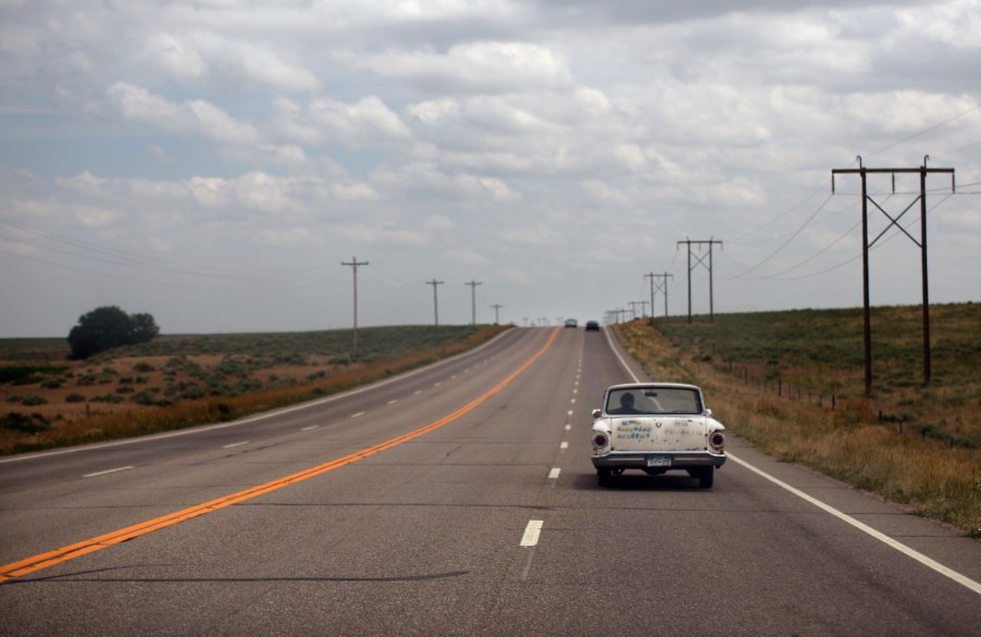 An old white station wagon car driving down an empty rural road with telephone poles and fields visible in the background.