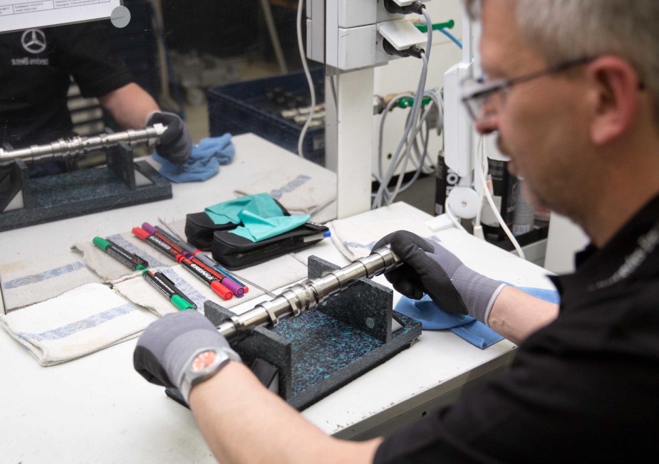 Shot over-the-shoulder as a Mercedes technician hones an advanced DOHC Camshaft, his workbench visible in the background.