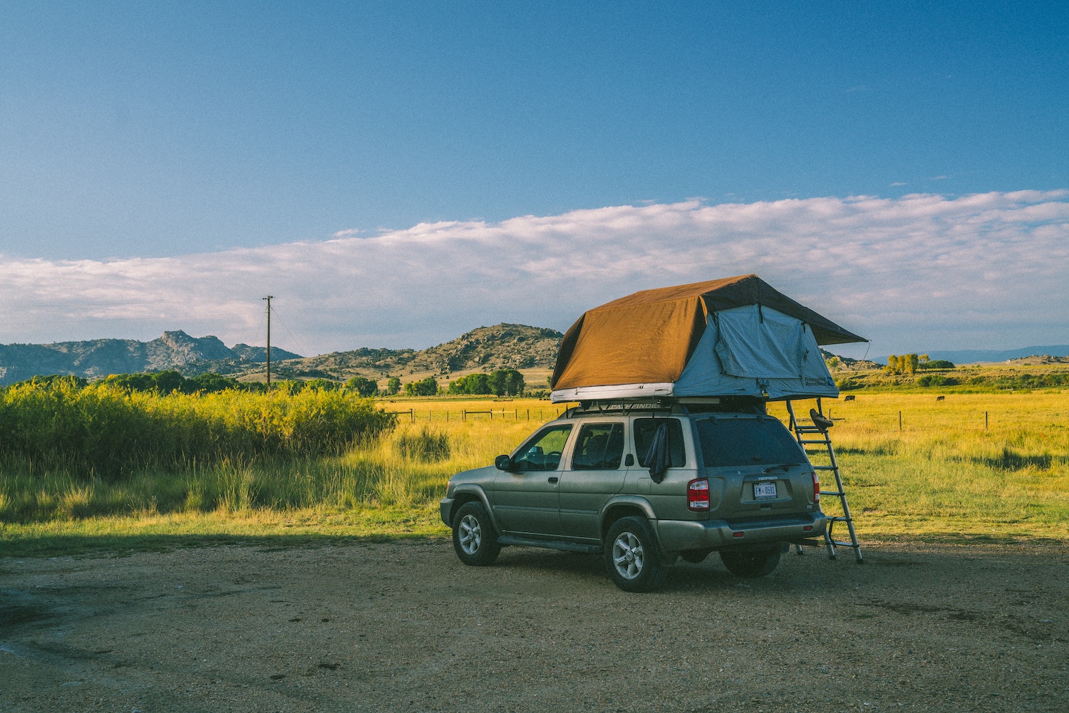 A brown Nissan Pathfinder parked in a rural lot with a soft-top rooftop tent on its rack, hills and power lines visible in the background.