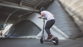 A man in a helmet races under a bridge on an electric kick scooter, a blurry stone wall visible in the background.