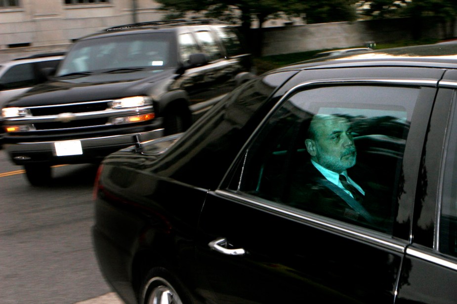 A man in a suit riding in the back seat of a Cadillac car with heavily tinted windows.