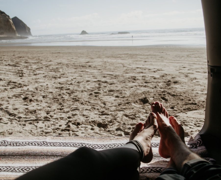 The view from a car parked on the beach, two people's feet visible in the foreground, the ocean in the background.