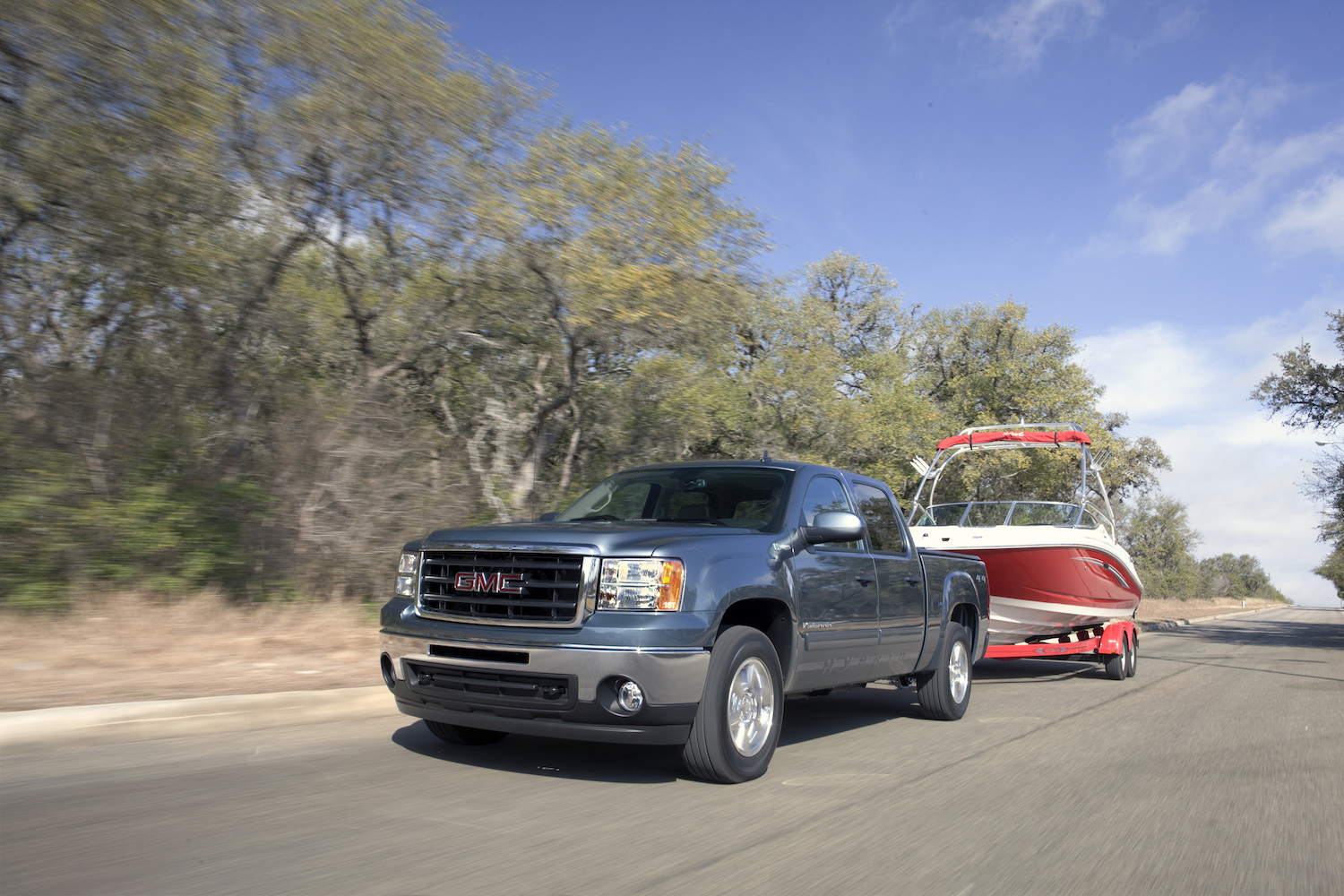 Promo photo of a GMC Sierra full-size hybrid pickup truck towing a boat down a rural road, trees visible in the background.