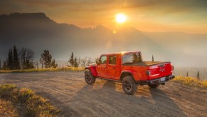 Promo photo of a red Jeep Gladiator 4WD truck navigating a dirt road, the sun setting behind a mountain range visible in the background.