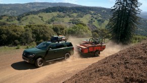 Green Ford Expedition Timberline edition full-size hybrid SUV towing a camping trailer up a dirt road, a mountain pass visible in the background.