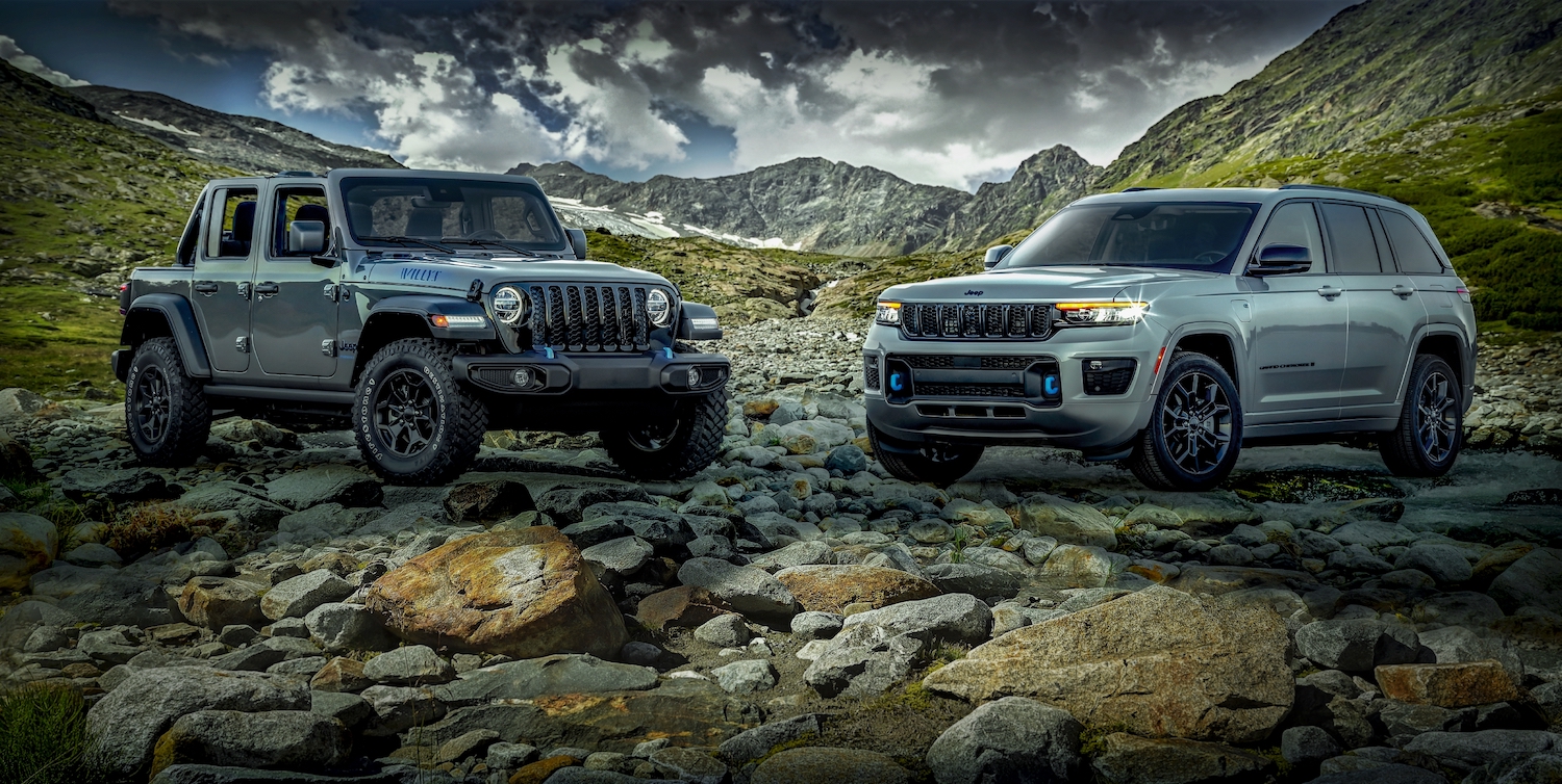 Two gray Jeep plug-in hybrid SUVs parked in a valley for a promo shot, mountains and clouds visible in the background.