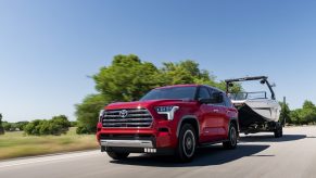 Red full-frame Toyota Sequoia hybrid SUV Towing a boat along a rural road, trees and sky visible behind it.