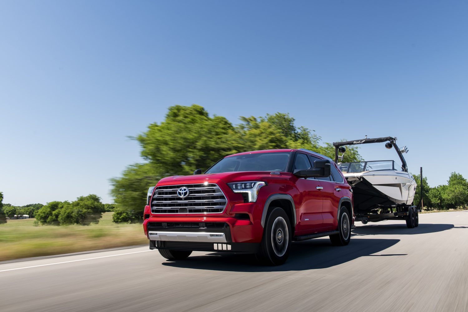 Red full-frame Toyota Sequoia hybrid SUV Towing a boat along a rural road, trees and sky visible behind it.