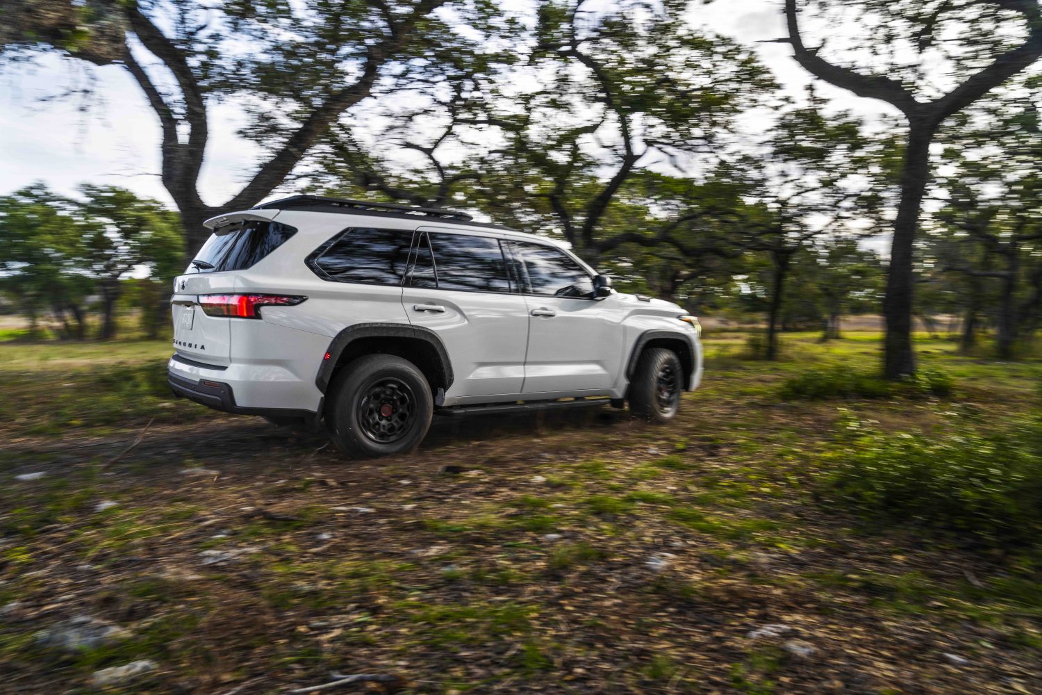 White Toyota Sequoia TRD Pro hybrid 3rd row SUV, driving through the woods, blurry trees visible in the background.