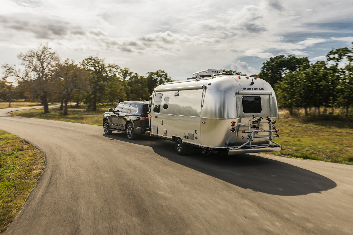 A 2023 Toyota Sequoia full-size SUV pulling an airstream camper trailer down a rural road.