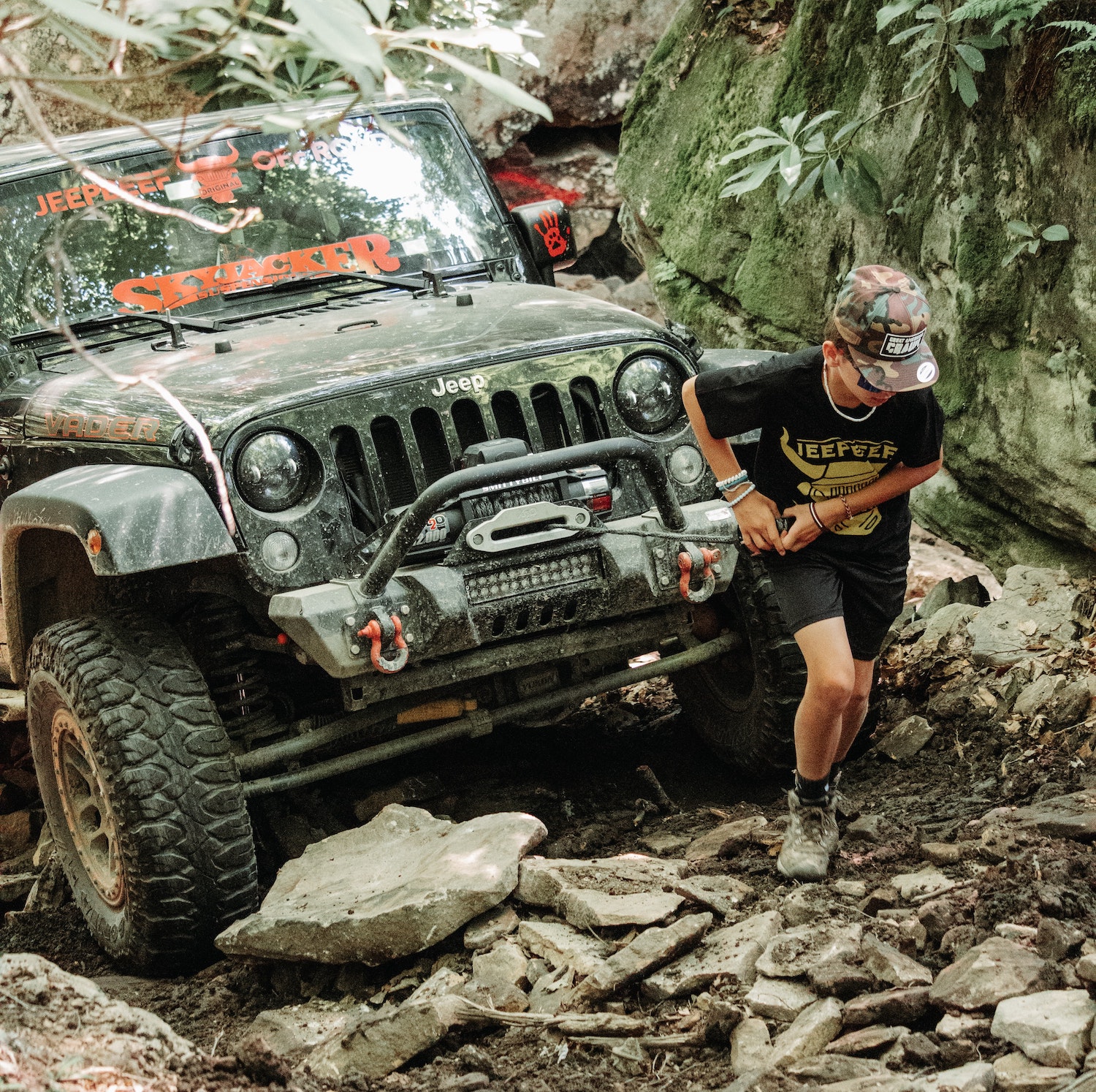 A young man deploys the winch from the front bumper of a 4x4 Jeep SUV, an off-road trail visible in the background.