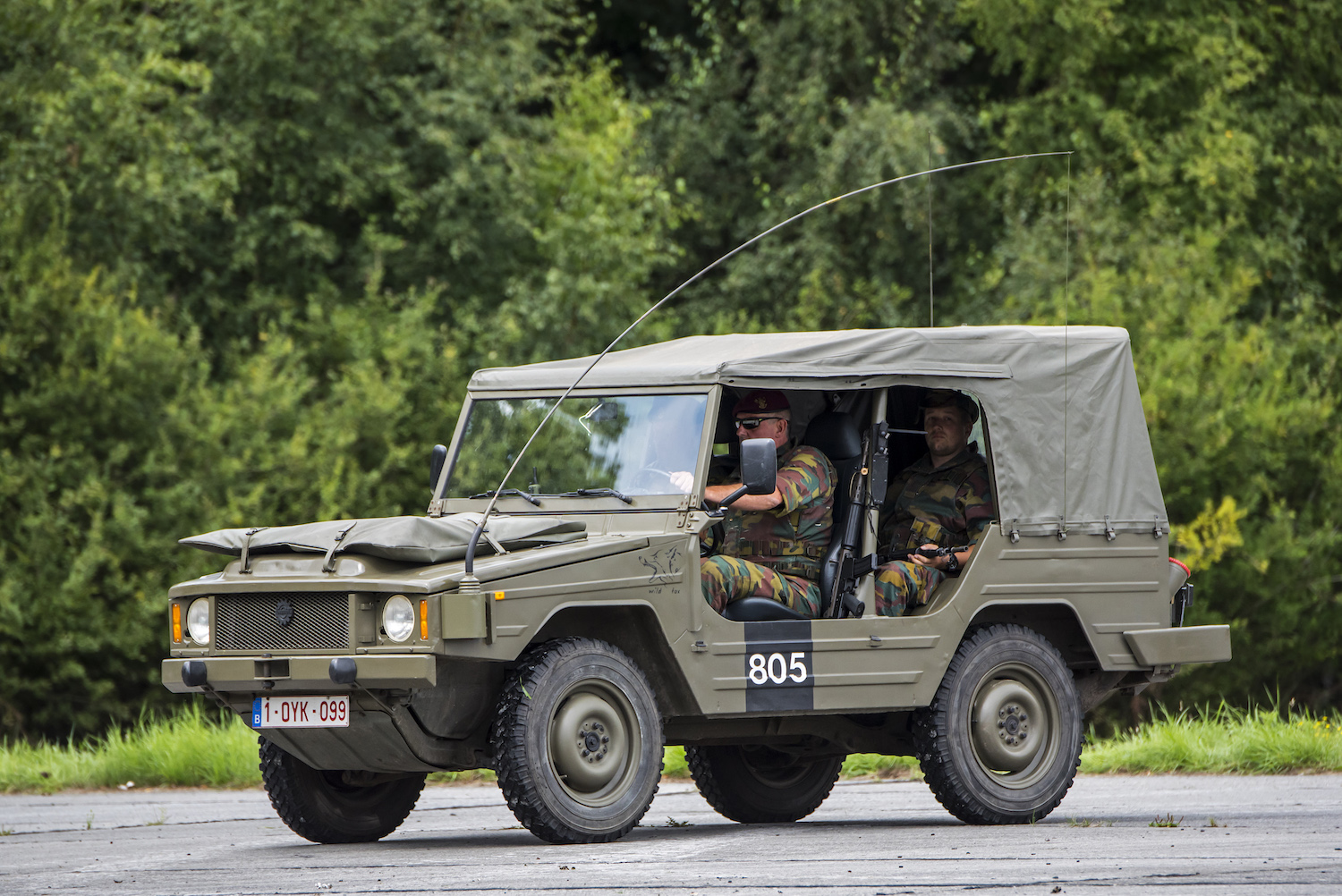 Soldier driving a military-spec Volkswagen type 183 Iltis on pavement, a wall of trees visible in the background.