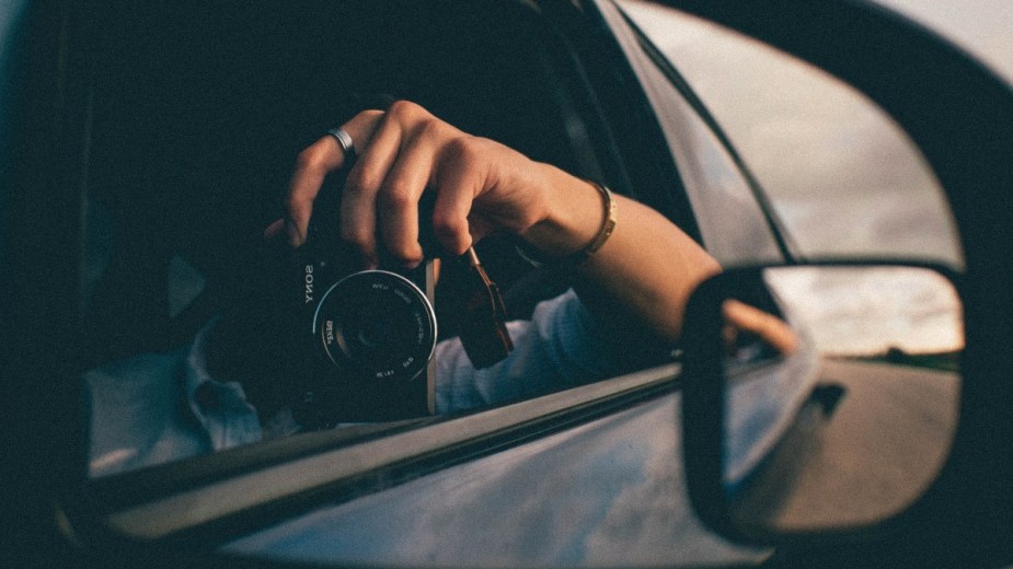 Person taking a selfie photo in the side-view mirror while driving a car, highlighting whether or not it's illegal