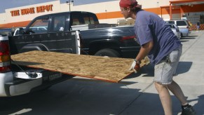 A man slides a sheet of plywood into the bed of a pickup truck, Home Depot visible behind him.