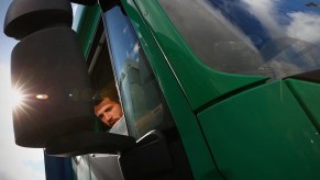 A truck driver, potentially with truck driver sun damage, in a green truck with a sunspot on the mirror.