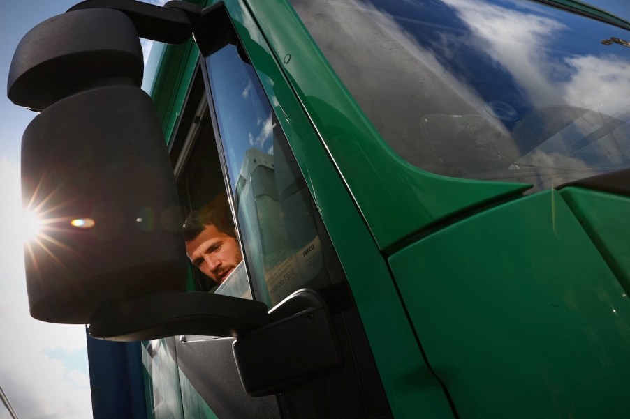 A truck driver, potentially with truck driver sun damage, in a green truck with a sunspot on the mirror.