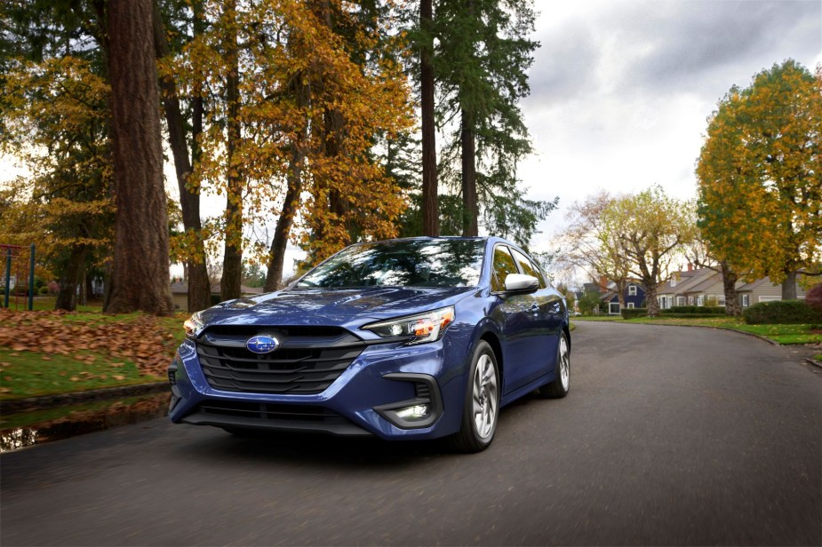 A blue 2023 Subaru Legacy midsize sedan model driving in a suburban neighborhood with autumn-colored trees