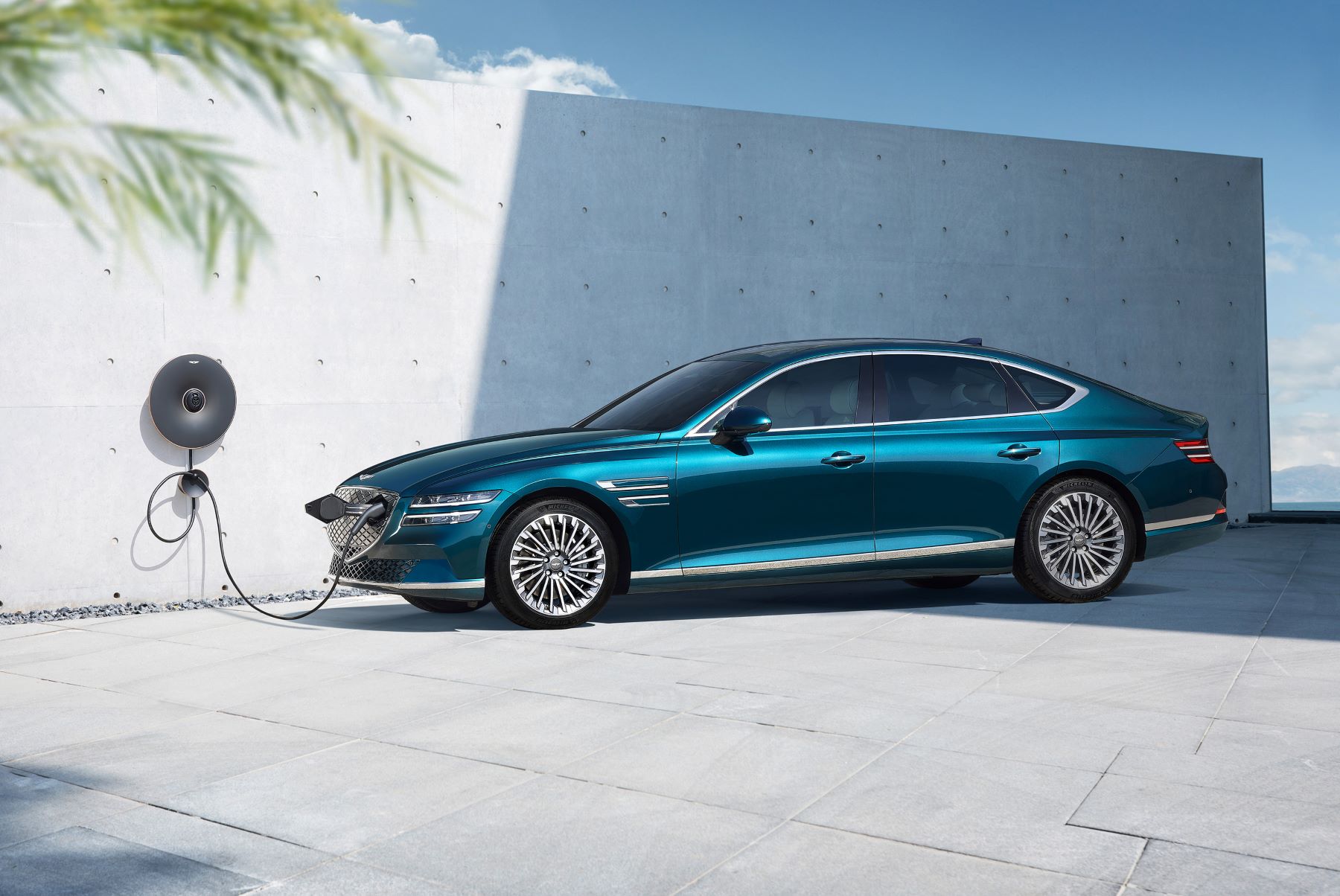 A teal Genesis Electrified G80 midsize EV sedan model plugged into a charging station