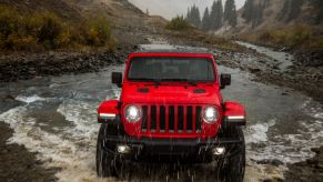 A red 2023 Jeep Wrangler off-road SUV driving through a muddy creek as it rains