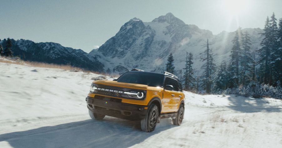 A yellow-gold 2022 Ford Bronco Sport driving off-road through a snowy field near a forest and mountains