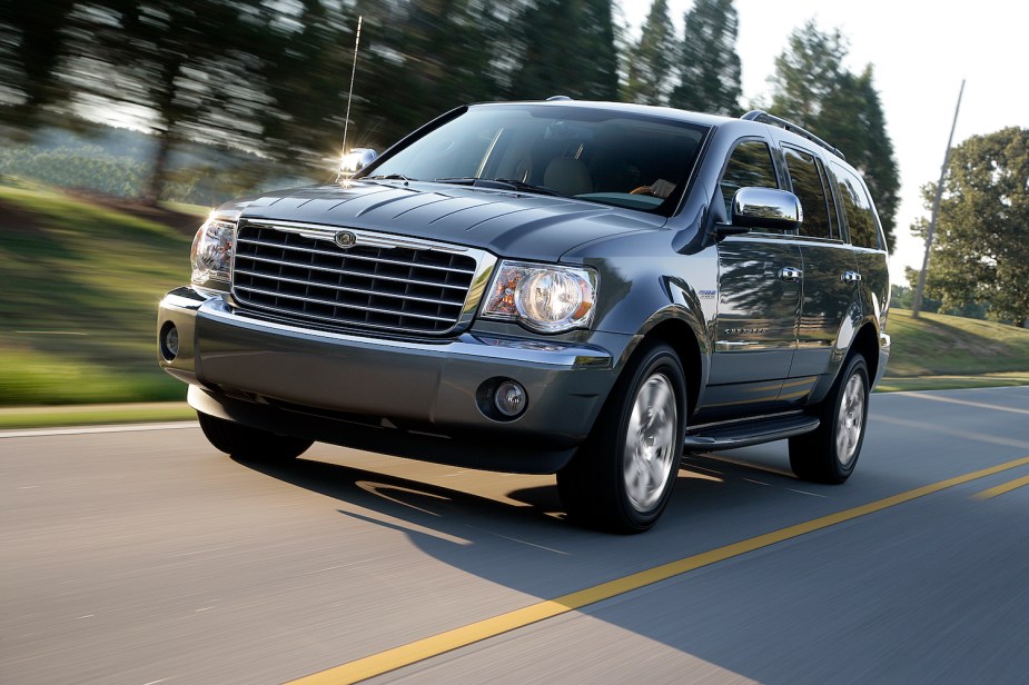 Promo photos of the Chrysler Aspen luxury hybrid full-size SUV driving down the road, trees visible in the background.