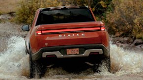 Red Rivian R1T electric pickup truck driving through a river, a field visible beyond it.