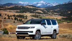 A white 2023 Jeep Grand Wagoneer luxury SUV parked off road, a row of snow-capped mountains visible in the background.