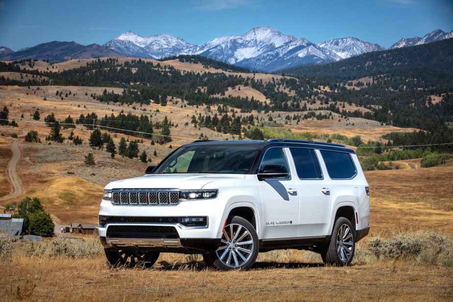 A white 2023 Jeep Grand Wagoneer luxury SUV parked off road, a row of snow-capped mountains visible in the background.