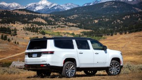 Promo photo of a white Grand Wagoneer SUV facing away from the camera, parked in front of a mountain range.